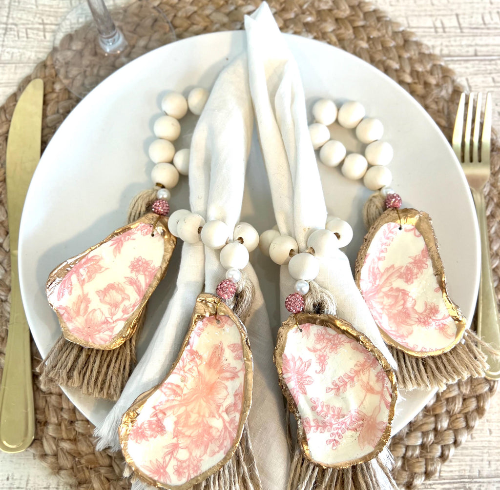 pink toile design oyster shell napkin rings with wood beads and gold leaf rim on a white plate with a white flower