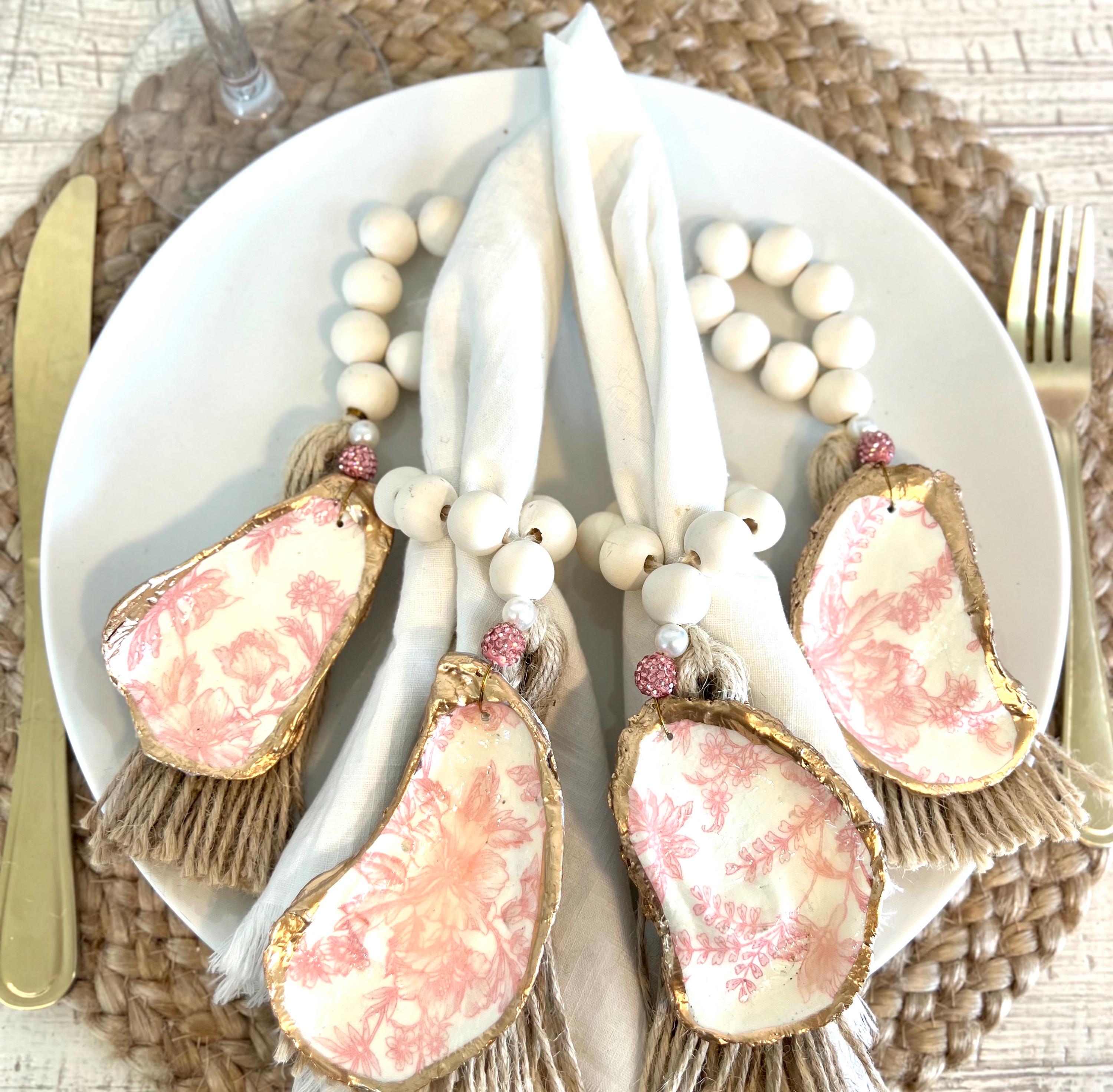 pink toile design oyster shell napkin rings with wood beads and gold leaf rim on a white plate with a white flower