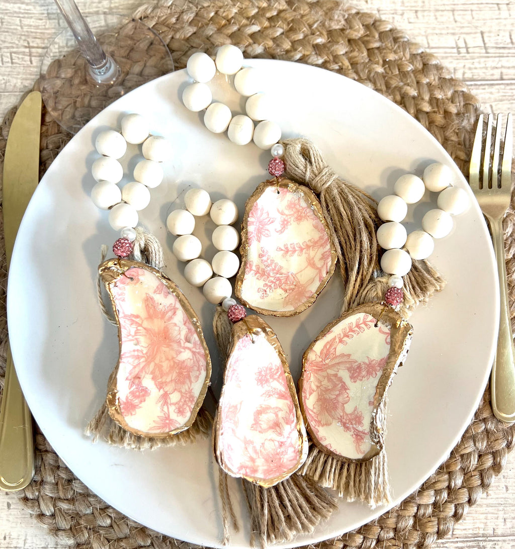 pink toile design oyster shell napkin rings with wood beads and gold leaf rim on a white plate with a white flower