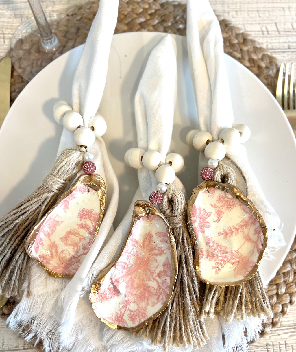 pink toile design oyster shell napkin rings with wood beads and gold leaf rim on a white plate with a white flower