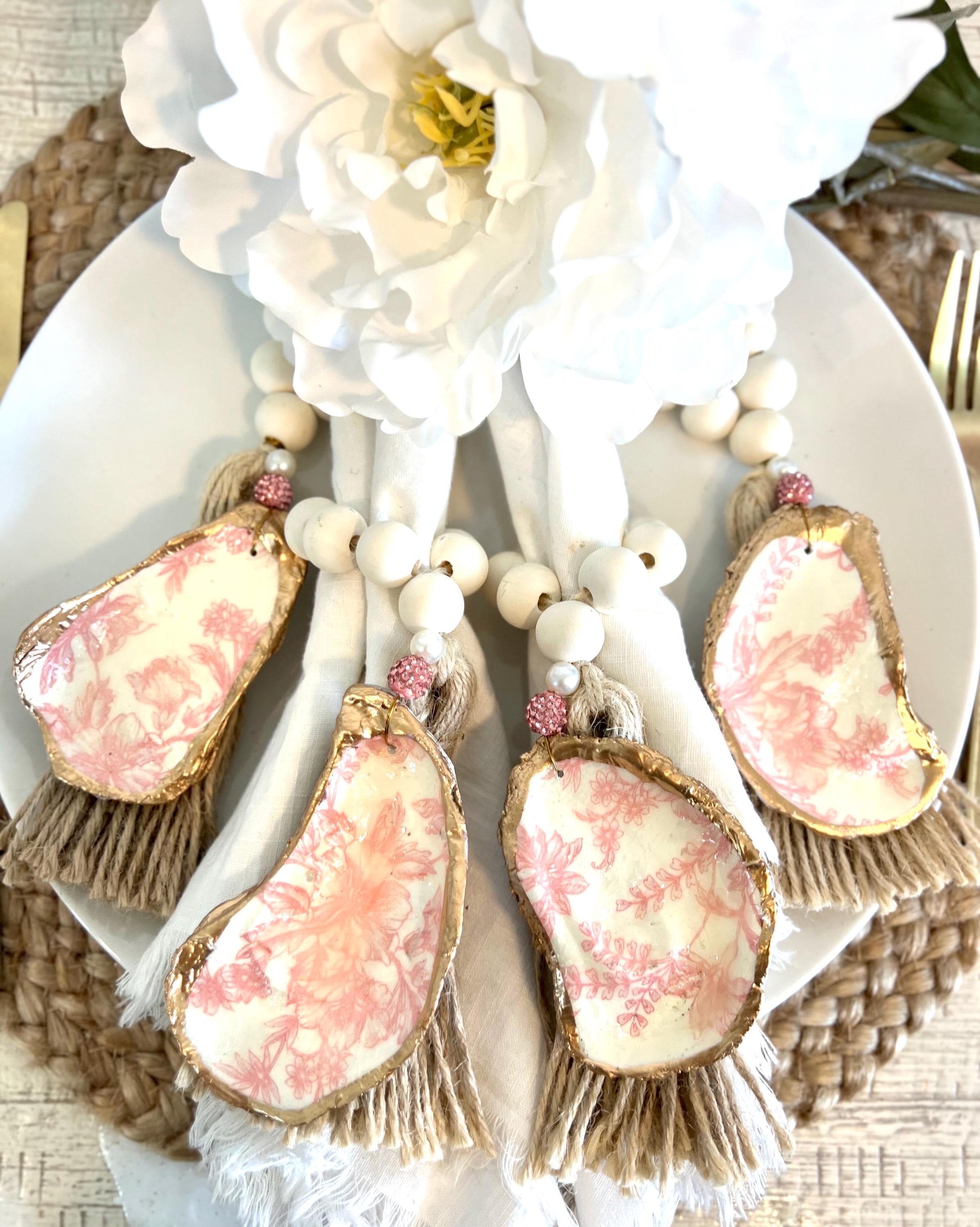 pink toile design oyster shell napkin rings with wood beads and gold leaf rim on a white plate with a white flower